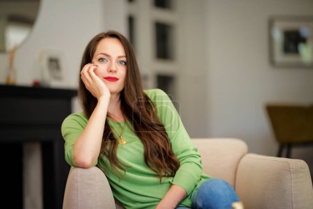Foto de Retrato de una hermosa mujer de pelo largo con ropa casual mientras se relaja en un sillón en casa. Atractiva joven hembra con lápiz labial rojo y suéter verde. - Imagen libre de derechos