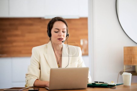 Photo for Customer service assistant wearing headset while sitting behind her computer and working in the call center. Attractive business woman wearing white blazer. - Royalty Free Image