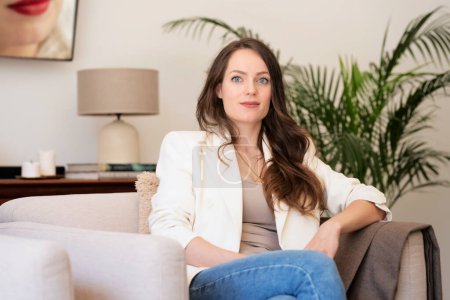 Téléchargez les photos : Portrait de belle femme aux cheveux longs portant un blazer blanc et un jean bleu et se relaxant dans un fauteuil à la maison. - en image libre de droit