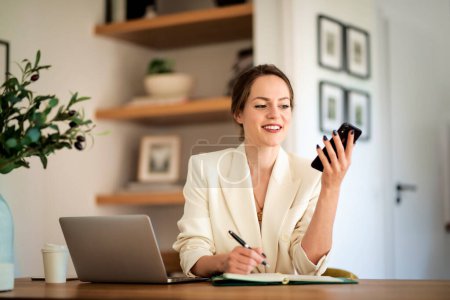 Photo for Attractive woman sitting at home and working on laptop. Business woman using smartphone and text messaging. Home office. - Royalty Free Image