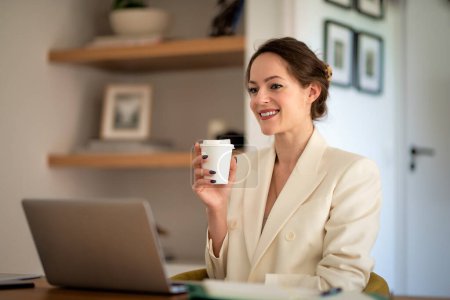 Foto de Atractiva joven mujer de negocios que usa el ordenador portátil mientras está sentada en el escritorio y tiene una videoconferencia en la oficina del hogar. - Imagen libre de derechos