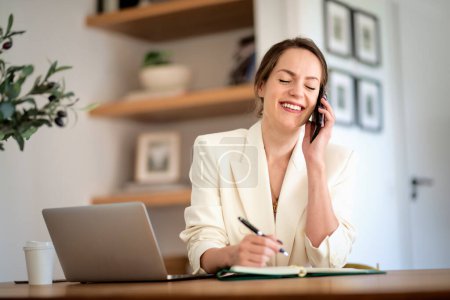 Foto de Mujer sonriente alegre sentada en casa y trabajando. Mujer de negocios usando laptop y haciendo una llamada. Oficina en casa. - Imagen libre de derechos