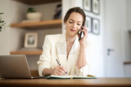 Photo for Attractive woman sitting at home and working. Business woman using laptop and making a call. Home office. - Royalty Free Image