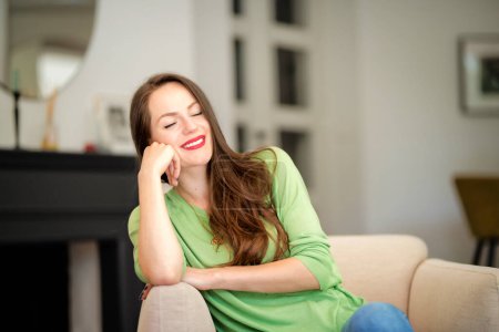 Photo for Portrait of beautiful long haired woman wearing casual clothes while relaxing in an armchair at home and daydreaming. Attractive young female wearing red lipstick and green sweater. - Royalty Free Image