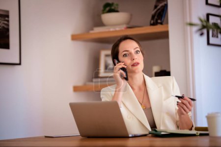 Foto de Mujer maravilla sentada en casa y trabajando. Mujer de negocios usando laptop y haciendo una llamada. Oficina en casa. - Imagen libre de derechos