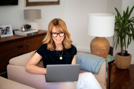 Téléchargez les photos : Portrait d'une jolie femme d'âge moyen assise à la maison et utilisant un ordinateur portable. Femme aux cheveux blonds portant des lunettes et une chemise noire. Bureau à domicile. - en image libre de droit