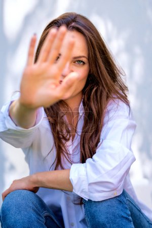 Photo for Portriat of a beautiful woman sitting in the sun, smiling and covering her face with her hands. Attractive female wearing white shirt and blue jeans. - Royalty Free Image