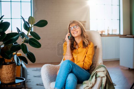 Photo for Portrait shot of happy mature woman talking with somebody on her mobile phone and relaxing at home in the armchair. - Royalty Free Image