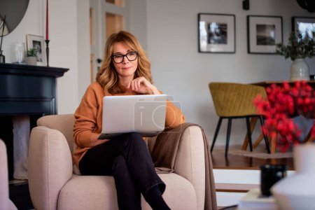 Foto de Mujer de mediana edad utilizando el ordenador portátil y tener videollamada mientras está sentado en casa y trabajando. Oficina en casa. - Imagen libre de derechos