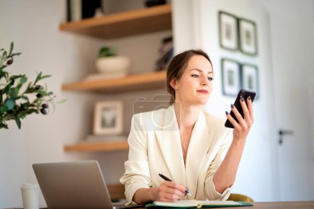 Photo for Attractive woman sitting at home and working on laptop. Business woman using smartphone and text messaging. Home office. - Royalty Free Image