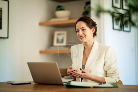 Photo for Attractive young woman using her laptop for video conferences while sitting at home. Confident businesswoman wearing white blazer. Home office. - Royalty Free Image