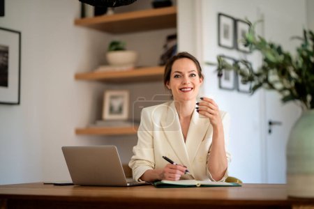 Photo for Attractive young business woman sitting at home and using laptop for work. Brunette haired woman wearing blazer and writing something. Home office. - Royalty Free Image