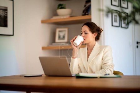 Photo for Attractive young business woman sitting at home and using laptop for work. Brunette haired woman wearing blazer and smiling. Home office. - Royalty Free Image