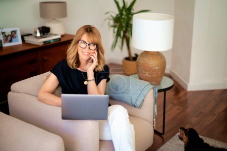 Photo for Portrait of attractive middle aged woman sitting at home and using laptop and cellphone. Blond haired female wearing eyeglasses and black shirt. Home office. - Royalty Free Image