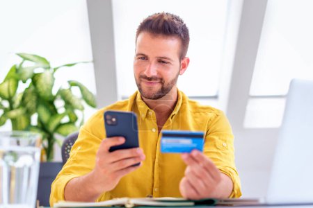 Téléchargez les photos : Prise de vue de l'homme tenant la carte de crédit et le téléphone portable dans ses mains tout en payant des factures avec carte de crédit. - en image libre de droit