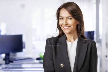 Foto de Atractiva mujer de negocios de mediana edad usando chaqueta y de pie en la oficina. Profesional hembra mirando a la cámara y sonriente alegre. - Imagen libre de derechos