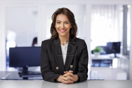 Foto de Atractiva mujer de negocios de mediana edad usando chaqueta y sentada en el escritorio de la oficina. Ejecutivo profesional de negocios mirando a la cámara y sonriendo. - Imagen libre de derechos