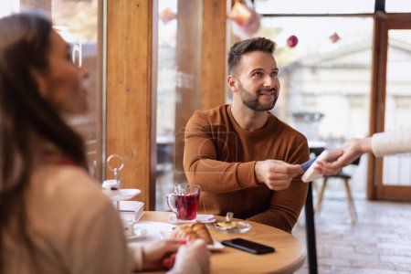 Foto de Primer plano de una pareja sentados juntos en una mesa en un café y el hombre pagando a la camarera con una tarjeta. Pago en línea. - Imagen libre de derechos