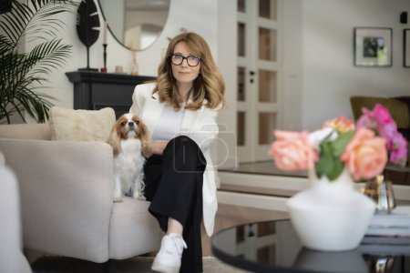 Photo for Happy attractive woman relaxing in an armchair at home. Mid aged female sitting at home on the sofa with her cute cavalier king charles puppy. - Royalty Free Image