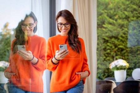Photo for Portrait shot of cheerful woman text messaging while standing at the window at home. Confident female wearing orange sweater and cheerful smiling. - Royalty Free Image