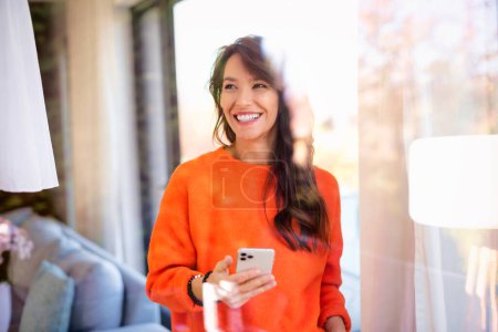 Photo for Portrait shot of cheerful woman text messaging while standing at the window at home. Confident female wearing orange sweater and cheerful smiling. - Royalty Free Image
