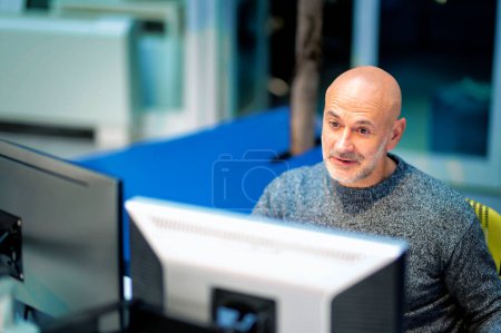 Photo for Smiling mid aged businessman sitting at office desk and using computers for work or having video call. - Royalty Free Image