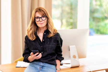 Photo for Close-up of a mid aged woman standing at desk and using mobile phone. Confident female wearing glasses and casual clothes and working from home. Home office. - Royalty Free Image
