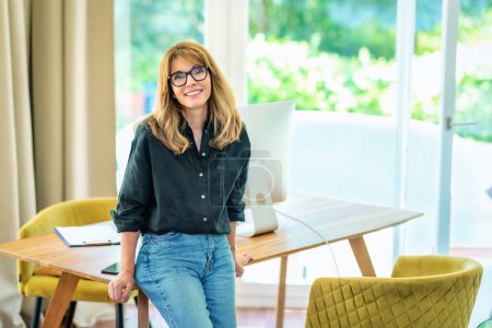 Photo for Portrait of mid aged businesswoman standing at desk while looking at camera and smiling. Confident female wearing black shirt and glasses. Home office. - Royalty Free Image