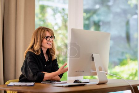 Photo for Mid aged woman sitting at desk and having web conference. Confident businesswoman using computer and working at home. Home office. - Royalty Free Image