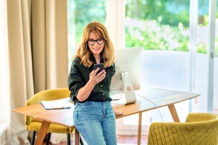 Photo for Shot of a mid aged woman standing at desk and using mobile phone. Confident female wearing glasses and casual clothes and text messaging. Home office. - Royalty Free Image