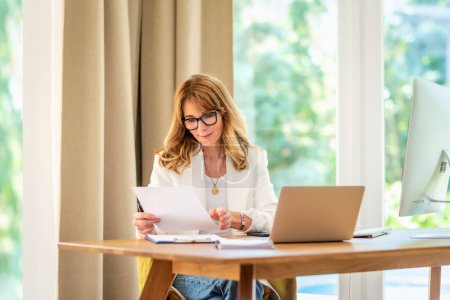 Photo for Shot of mid aged woman sitting at desk and using laptop for work. Confident businesswoman doing some paperwork while working from home. Home office. - Royalty Free Image