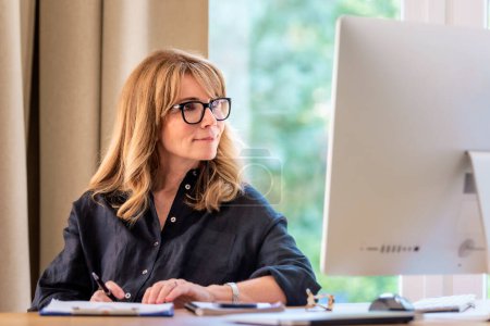 Photo for Close-up of mid aged woman sitting at desk and using computer for work. Confident businesswoman doing some paperwork while working from home. Home office. - Royalty Free Image