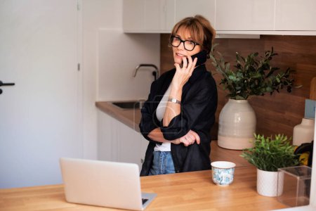 Photo for Shot of middle aged woman using laptop and having a call while standing in the kitchen at home. Home office. - Royalty Free Image