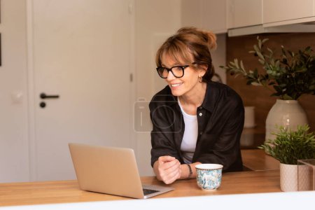Photo for Shot of middle aged woman standing in the kitchen at home. and using laptop. Confident female having video call. Home office. - Royalty Free Image