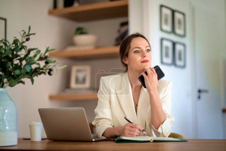 Téléchargez les photos : Jolie jeune femme d'affaires assise à la maison et utilisant un ordinateur portable pour le travail. Femme aux cheveux bruns portant un blazer et écrivant quelque chose. Bureau à domicile. - en image libre de droit