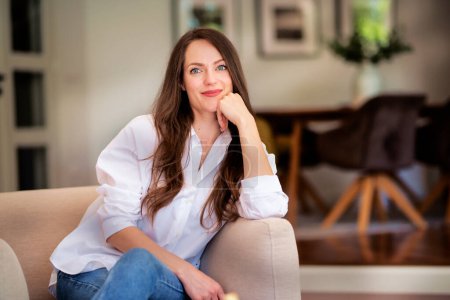 Foto de Hermosa mujer de pelo largo con camisa blanca y relajante en un sillón en casa. Atractiva hembra mirando pensativamente. - Imagen libre de derechos