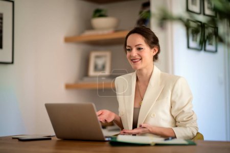 Téléchargez les photos : Jolie jeune femme d'affaires assise à la maison et utilisant un ordinateur portable pour le travail. Femme aux cheveux bruns portant un blazer et ayant une vidéoconférence. Bureau à domicile. - en image libre de droit