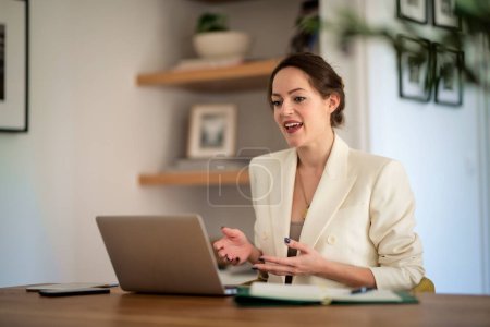 Téléchargez les photos : Jolie jeune femme d'affaires assise à la maison et utilisant un ordinateur portable pour le travail. Femme aux cheveux bruns portant un blazer et ayant un appel vidéo. Bureau à domicile. - en image libre de droit