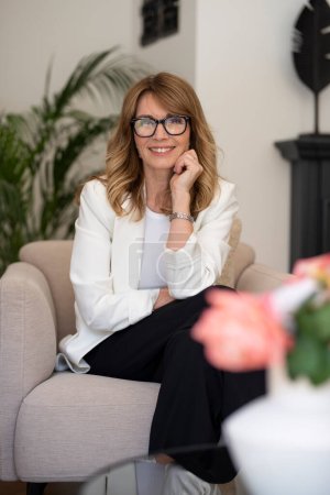 Photo for Happy attractive woman relaxing in an armchair at home. Blond haired female wearing eyeglasses and white blazer. - Royalty Free Image
