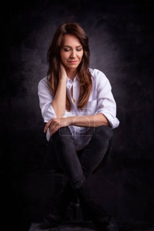 Foto de Atractiva mujer de mediana edad con camisa blanca y jeans negros mientras se sienta en un fondo oscuro aislado. Copiar espacio. Captura de estudio. - Imagen libre de derechos
