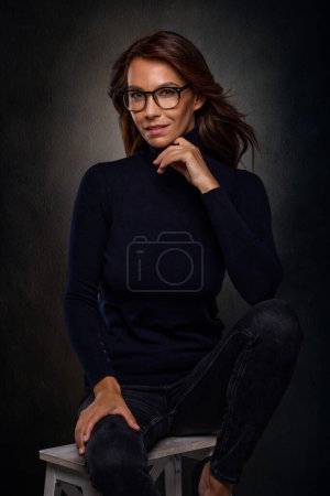 Téléchargez les photos : Jolie femme d'âge moyen portant un pull à col roulé et des lunettes alors qu'elle était assise sur un tabouret sur un fond sombre isolé. Espace de copie. Plan studio. - en image libre de droit