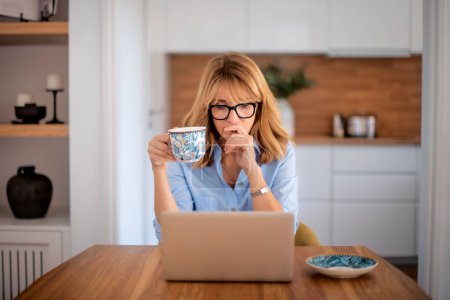 Téléchargez les photos : Pensant femme d'âge moyen en utilisant un ordinateur portable et boire du thé tout en travaillant à la maison. Femme confiante assise à table à la cuisine. Bureau à domicile. - en image libre de droit