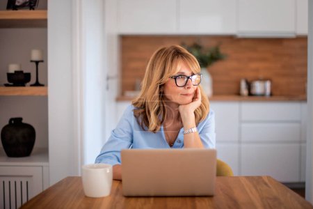 Photo for Smiling middle aged woman using laptop and working from home. Confident female deep in thought  while sitting at table at the kitchen. Home office. - Royalty Free Image