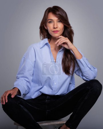 Téléchargez les photos : Portrait en studio d'une jolie femme portant une chemise et souriant assis sur un fond gris isolé. Espace de copie. - en image libre de droit