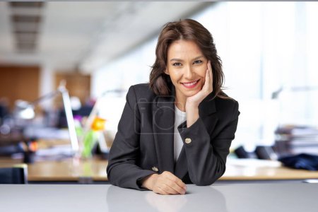 Téléchargez les photos : Portrait d'une jolie femme d'affaires assise au bureau. Femme professionnelle d'âge moyen portant un blazer noir et regardant la caméra. - en image libre de droit