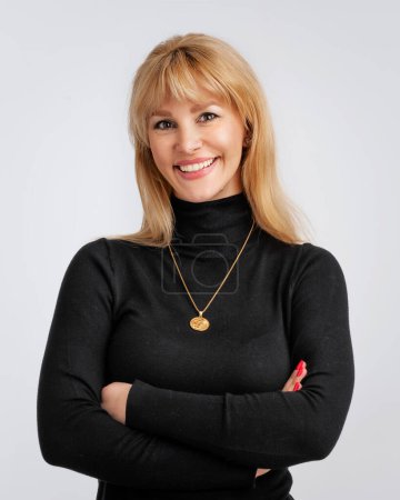 Foto de Una atractiva mujer rubia vistiendo suéter negro. Mujer sonriente de pie con los brazos cruzados. Ella está contra el fondo gris. Copiar espacio. - Imagen libre de derechos