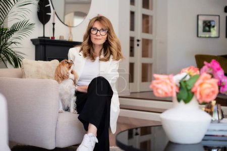 Photo for Attractive woman relaxing in an armchair at home with her cute cavalier king charles puppy. Mid aged female wearing eyewear and white blazer. Full length shot. - Royalty Free Image