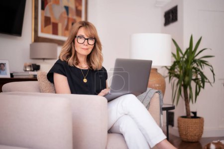 Foto de Atractiva mujer de mediana edad sentada en casa y usando un portátil. Mujer de pelo rubio con anteojos y camisa negra. Oficina en casa. - Imagen libre de derechos