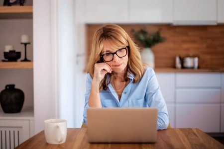 Téléchargez les photos : Une femme d'âge moyen avec un ordinateur portable travaillant à la maison. Une femme aux cheveux blonds est assise à table et travaille sur son ordinateur portable. Bureau à domicile. - en image libre de droit