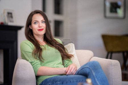 Foto de Una mujer sonriente sentada en un sillón en una casa moderna y relajante. Morena peluda mujer usando ropa casual. - Imagen libre de derechos
